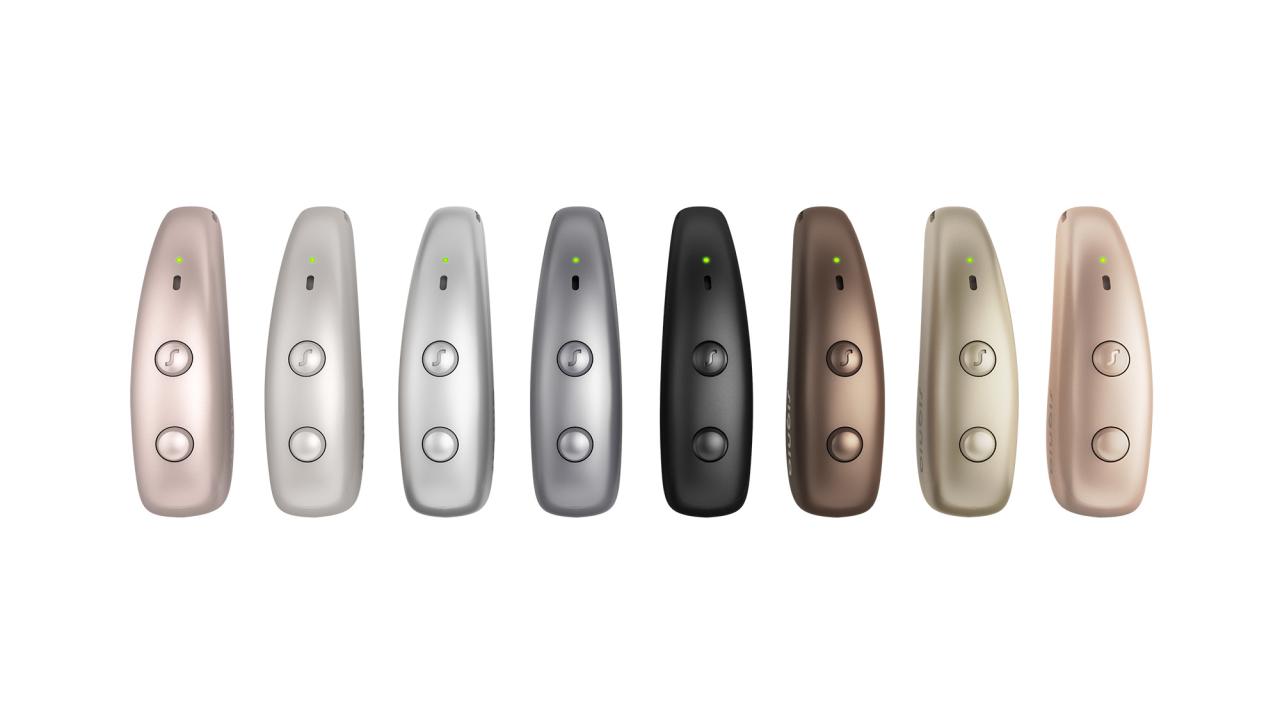 Seven sleek, metallic Signia Pure Charge&Go Max hearing aids in various colors (rose gold, silver grey, titanium grey, black, chestnut brown, honey blonde, beige, and dark champagne) arranged in a row against a solid white background.