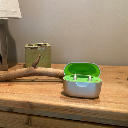 a green and white case of hearing aids on a wooden table. A stick, lamp, and pot are in the background