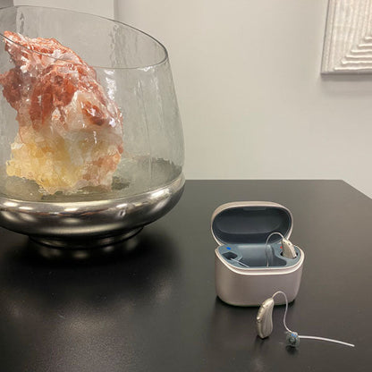 Hearing aid case sitting on table with 1 hearing aid inside case and 1 sitting on table. 