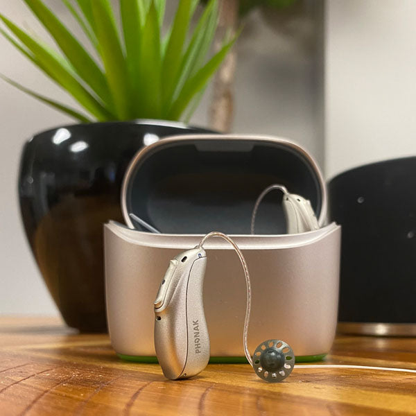 Hearing aid case sitting on table with 1 hearing aid inside case and 1 sitting on table. 