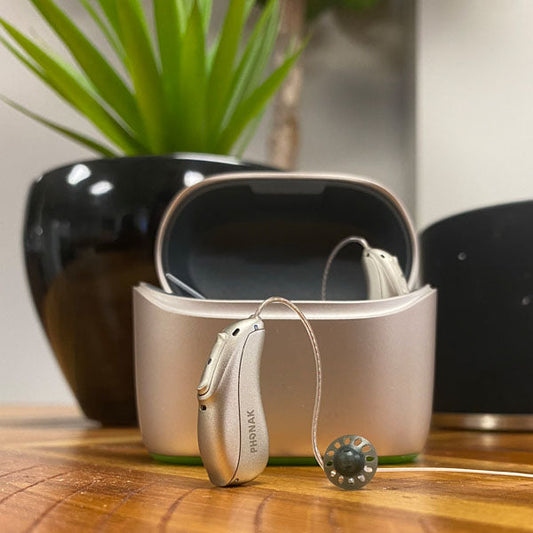 Hearing aid case sitting on table with 1 hearing aid inside case and 1 sitting on table. 